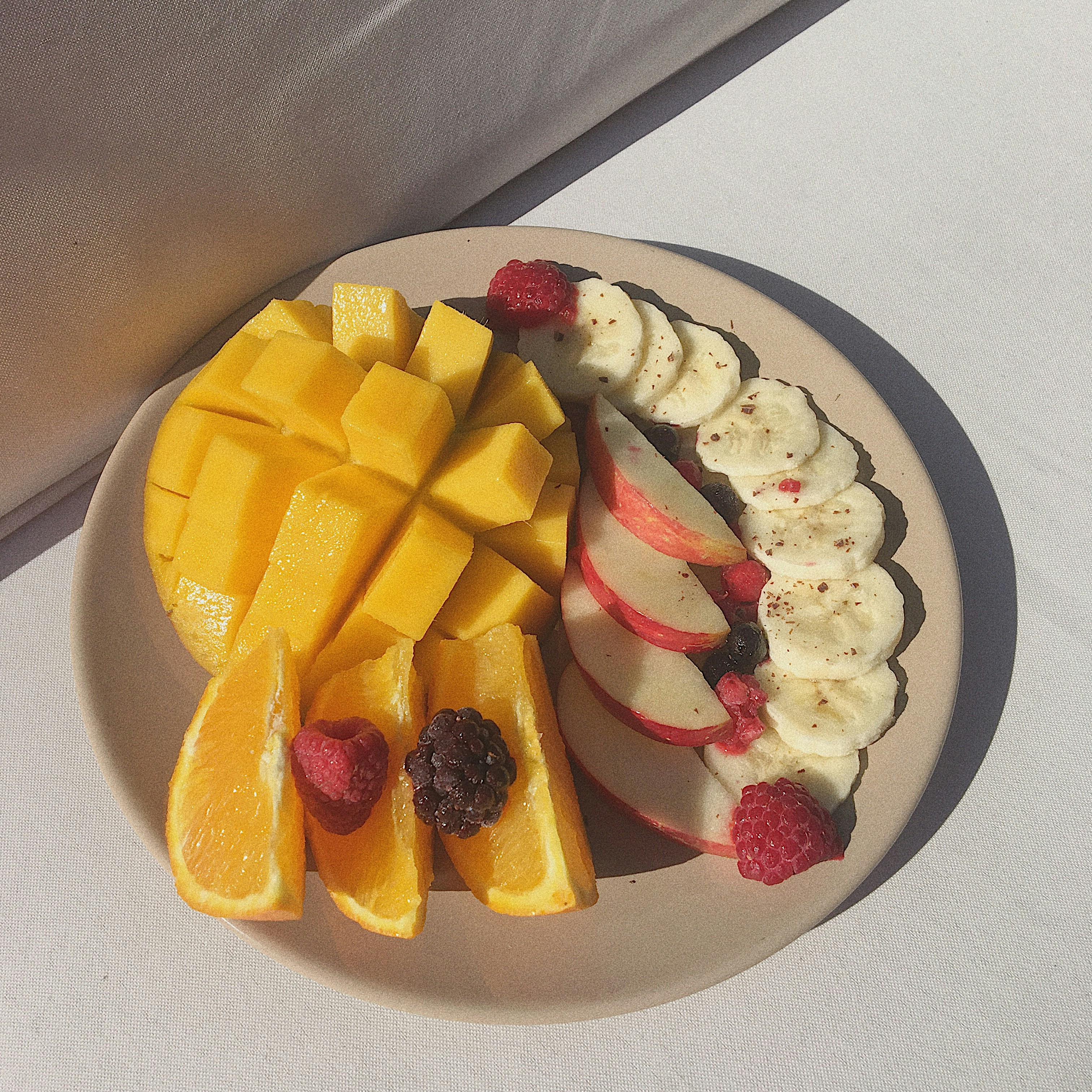 A plate of sliced tropical fruit — mango, banana, and berries — in morning light