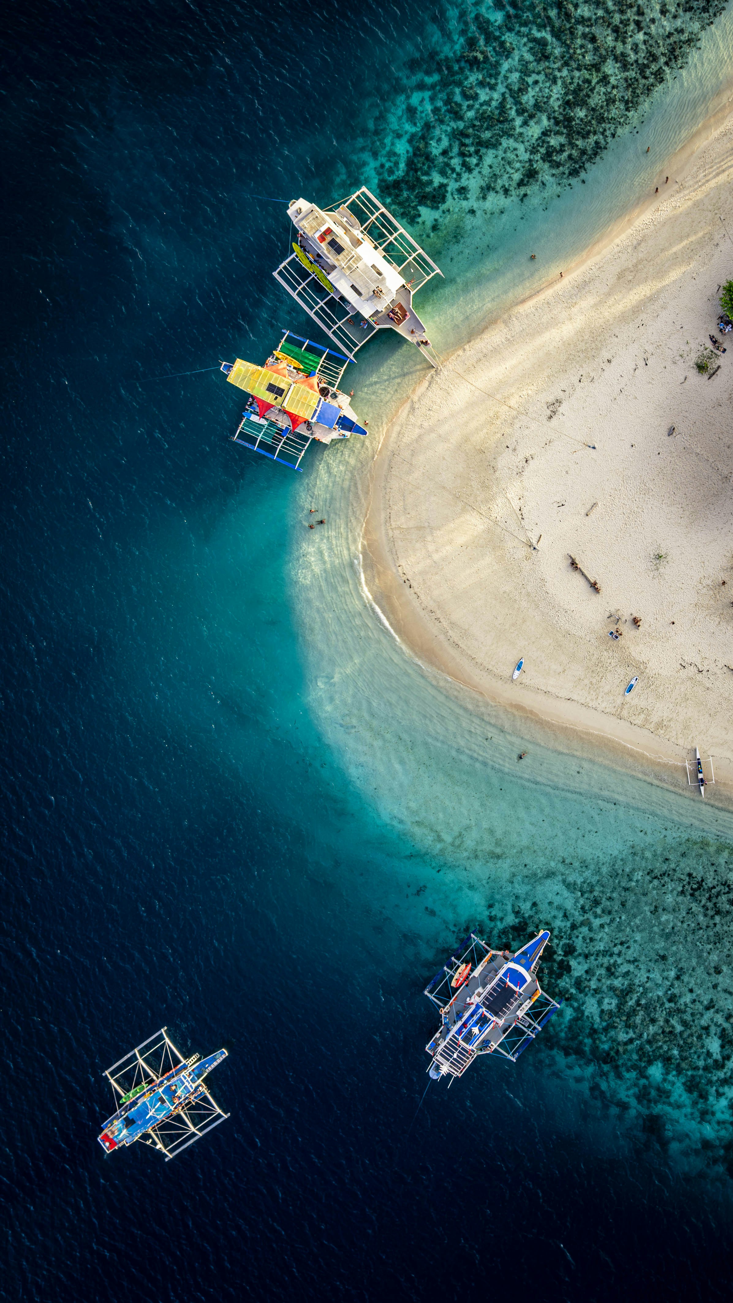A white sandbar with outrigger boats anchored in shallow turquoise water, seen from above
